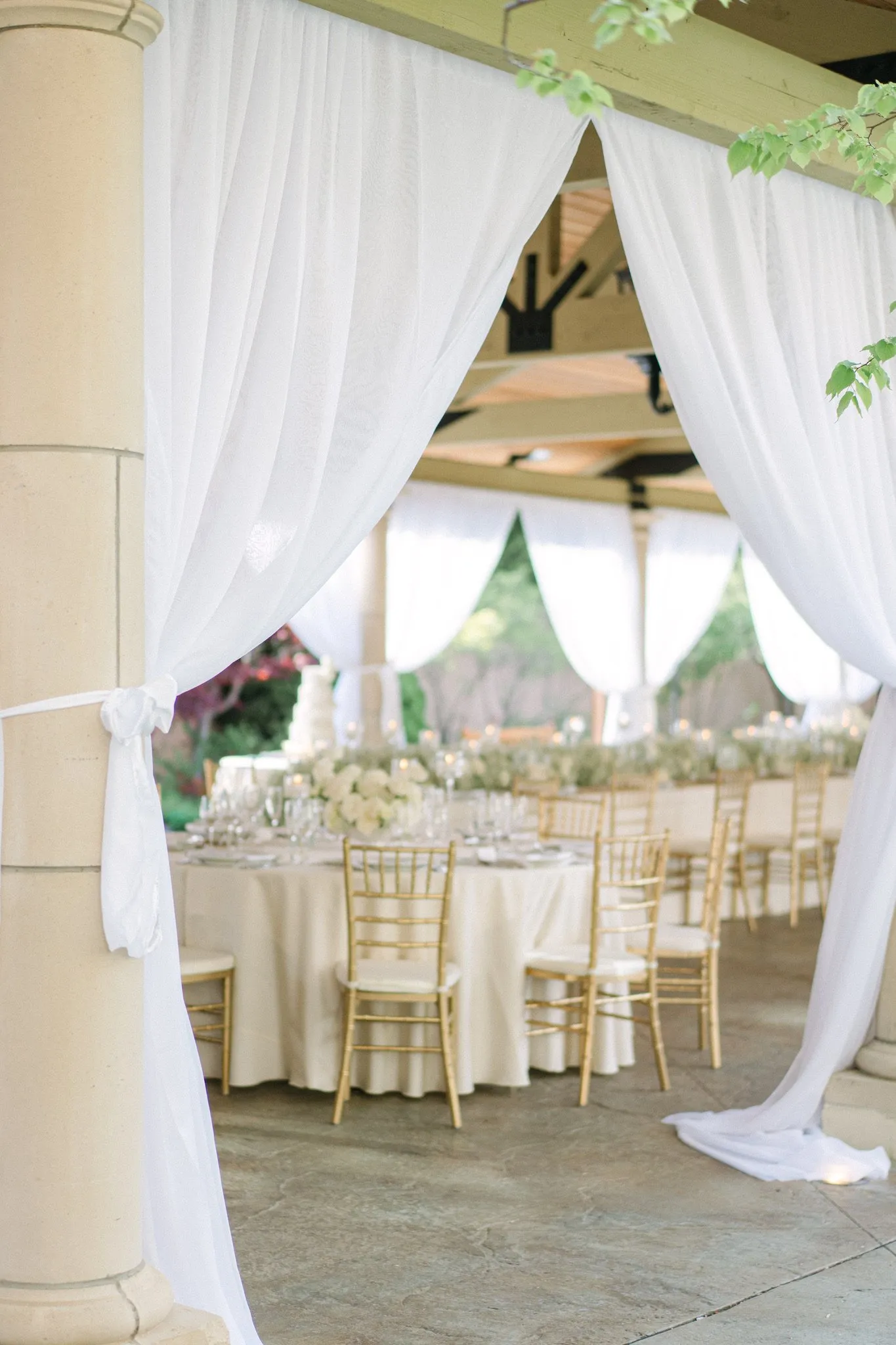 Outdoor event with white draping, floral arrangements, and gold chairs